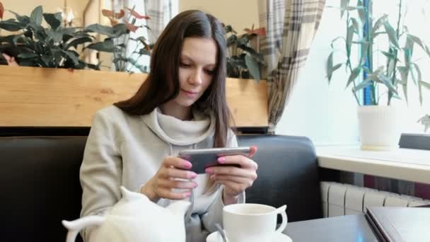Νεαρή γυναίκα μελαχρινή εικόνες τσαγιέρα και Κυπέλλου, σε ένα καφενείο στο κινητό της. — Αρχείο Βίντεο