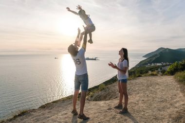 Baba oğlu havaya atar ve anne onlara dağda deniz manzarası ile bakıyor. Aile gezisi.