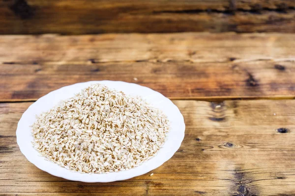 Close-up dry oat flakes in white plate in a wood background. Farm-fresh product. Side view. — Stock Photo, Image