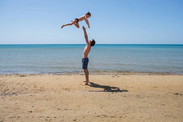 Joven padre feliz sosteniendo en sus brazos pequeño hijo ponerlo en la playa . — Foto de Stock
