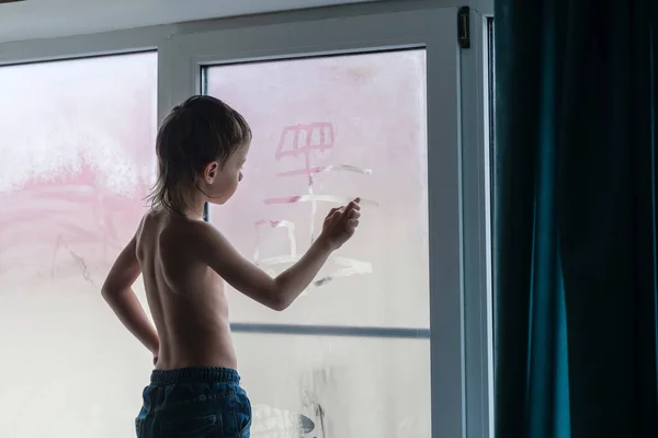 Шестилетний мальчик в джинсах рисует лодку на туманном окне, стоящем на подоконнике дома . — стоковое фото