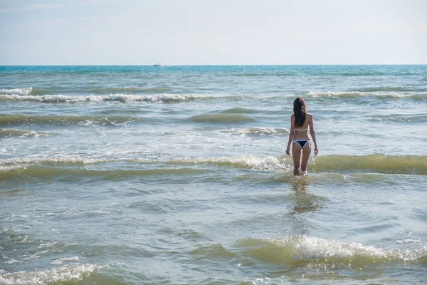 Mujer morena irreconocible con el pelo largo camina hacia el mar con olas. Vista trasera . — Foto de Stock