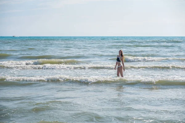 Bruenette kobieta z długimi włosami w niebieskie bikini spaceru w wodzie morskiej z falami. Widok z boku. — Zdjęcie stockowe