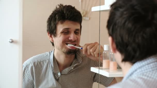 Müder, schläfriger Mann mit Kater, der gerade aufgewacht ist und seine Zähne putzt, blickt auf sein Spiegelbild im Spiegel. — Stockvideo