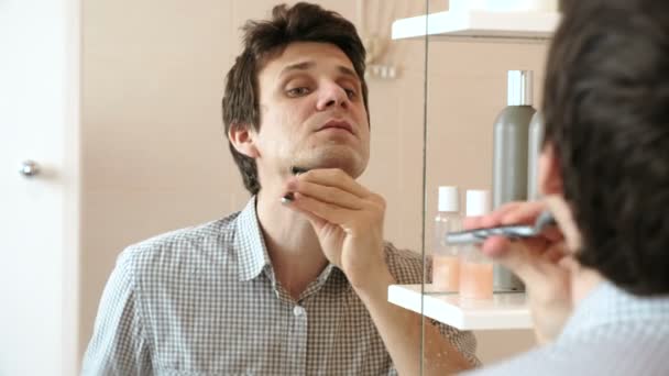 Mann rasiert sich beim Blick in den Spiegel. brünett im karierten hellen Hemd. — Stockvideo