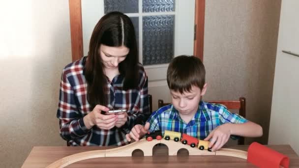 Jugando juntos. Mamá mira el teléfono y el hijo está jugando un ferrocarril de madera con tren, vagones y túnel sentado a la mesa . — Vídeos de Stock