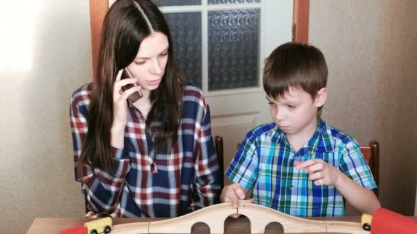 Samenspelen. Moeder praat haar telefoon en zoon is een houten spoorlijn met trein, wagons en tunnel zitten aan de tafel spelen. — Stockvideo