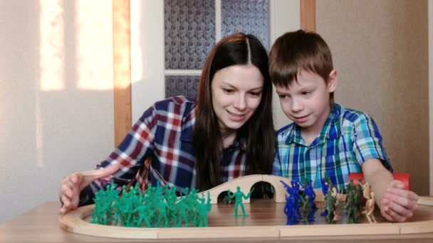 Zusammen zu spielen. Mutter und Sohn spielen eine hölzerne Eisenbahn mit Zug, Waggons und Tunnel mit Plastiksoldaten am Tisch. — Stockvideo
