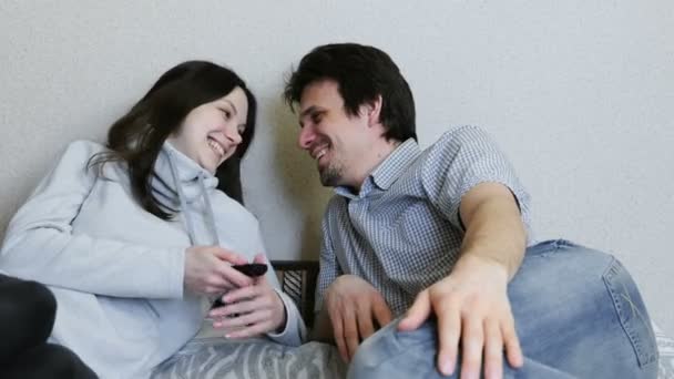 Парочка смотрит телевизор, лежащий на диване. Мужчина и женщина смотрят друг на друга и улыбаются. . — стоковое видео