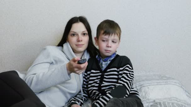 Viendo la tele tirada en el sofá. Mamá e hijo ven la televisión juntos y sonríen . — Vídeos de Stock