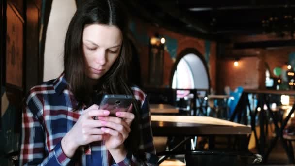 Mulher perturbada envia uma mensagem ou usar a internet no telefone sentado em um café. Vestido com uma camisa xadrez . — Vídeo de Stock