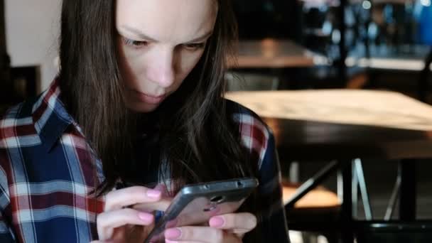 Upprörd kvinna skickar ett meddelande eller använda internet i telefonen sitter på ett café. Klädd i en rutig skjorta. — Stockvideo