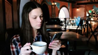 Büyük bir bardaktan çay içme ve seyir telefon Kareli gömlekli esmer kadın.