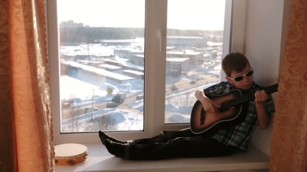 Tocar un instrumento musical. Niño en gafas de sol toca la guitarra sentado en el alféizar . — Vídeos de Stock