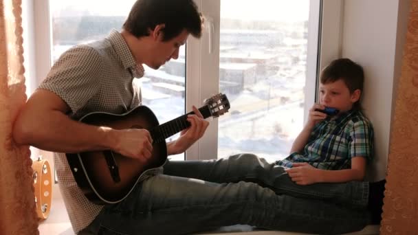 A tocar um instrumento musical. Pai está tocando guitarra e filho está tocando órgão da boca sentado no peitoril da janela . — Vídeo de Stock