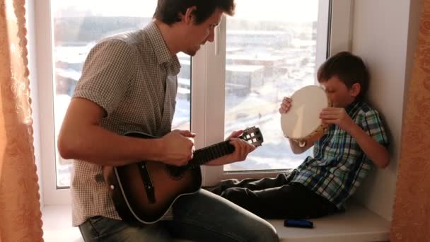 Suonare uno strumento musicale. Papà sta suonando la chitarra e il figlio suona il tamburello seduto nel davanzale della finestra . — Video Stock