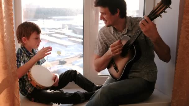 Playing a musical instrument. Dad is playing the guitar and son is playing tambourine sitting in windowsill. — Stock Video