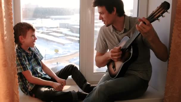 Jouer d'un instrument de musique. Papa joue de la guitare et le fils joue du tambourin assis dans le rebord de la fenêtre . — Video