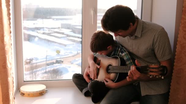 A tocar um instrumento musical. Pai ensina seu filho a tocar guitarra, sentado no peitoril da janela . — Vídeo de Stock