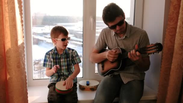 サングラスで楽器を演奏します。パパがギターを弾いているし、息子が窓辺に座ってドラムを再生. — ストック動画
