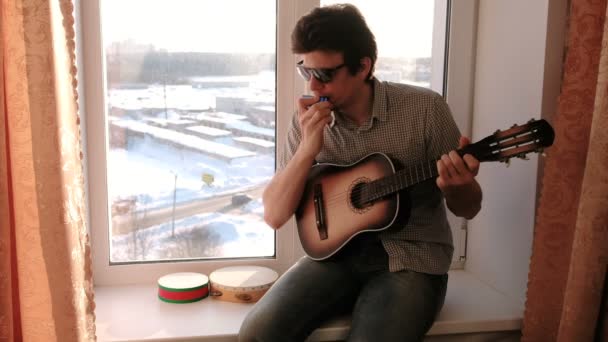 Playing a musical instrument. Man is playing the mouth organ sitting in windowsill — Stock Video