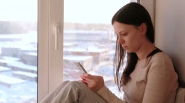 Sıkılmış kadın bir şey Internet üzerinde bir pencere üzerinde otururken cep telefonu izliyor.