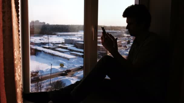 Silhouette d'un homme avec téléphone portable assis sur le rebord de la fenêtre . — Video