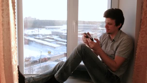 Man playing games in his mobile phone and laughs while sitting on a windowsill. — Stock Video