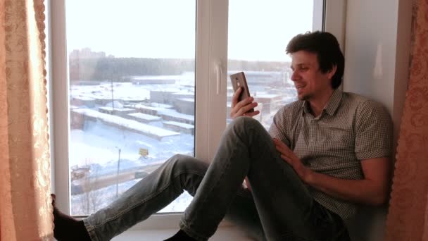 Homme heureux parlant sur la communication vidéo sur un téléphone mobile assis sur un rebord de fenêtre . — Video