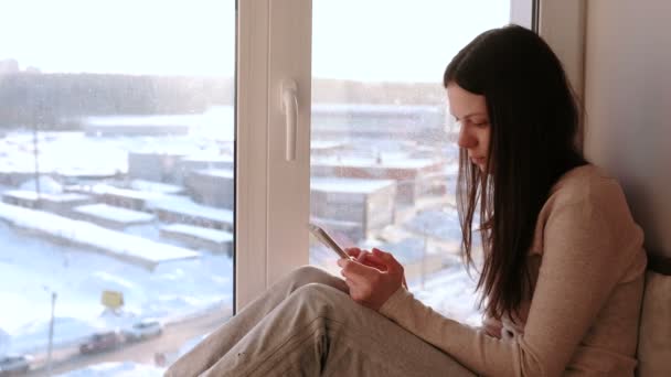 Mujer aburrida ve algo en Internet en su teléfono móvil mientras está sentada en un alféizar de la ventana . — Vídeos de Stock