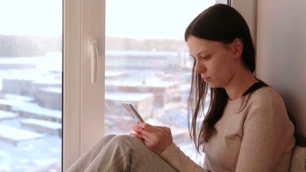 Mujer aburrida ve algo en Internet en su teléfono móvil mientras está sentada en un alféizar de la ventana . — Vídeos de Stock