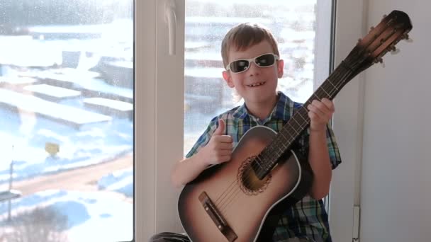Грає на музичному інструменті. Хлопчик грає на гітарі і співає, сидячи на підвіконні . — стокове відео