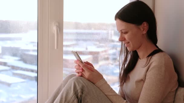 Mujer aburrida ve algo en Internet en su teléfono móvil y sonríe mientras se sienta en un alféizar de la ventana — Vídeos de Stock