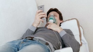 Nebulizatör ve astım tedavisi için kullanın. Genç adam kanepede yatan ve hareket eden telefon içinde sohbet inhaler maskesi aracılığıyla teneffüs edilmesi. Yan görünüm.