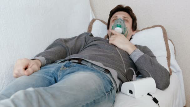 Use nebulizador e inhalador para el tratamiento. Hombre joven inhalando a través de la máscara del inhalador acostado en el sofá. Vista frontal — Vídeo de stock