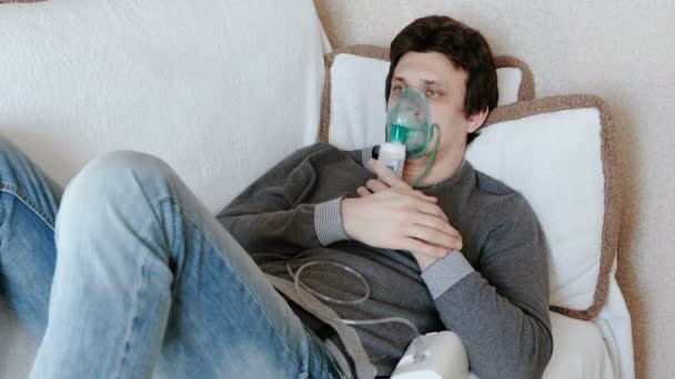 Use nebulizador e inhalador para el tratamiento. Hombre joven inhalando a través de la máscara del inhalador acostado en el sofá. Vista frontal — Vídeo de stock