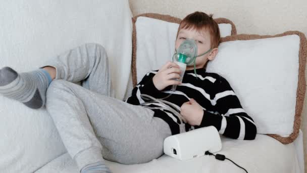 Использование распылителя и ингалятора для лечения. Мальчик вдыхает через маску ингалятора, лежащую на диване . — стоковое видео
