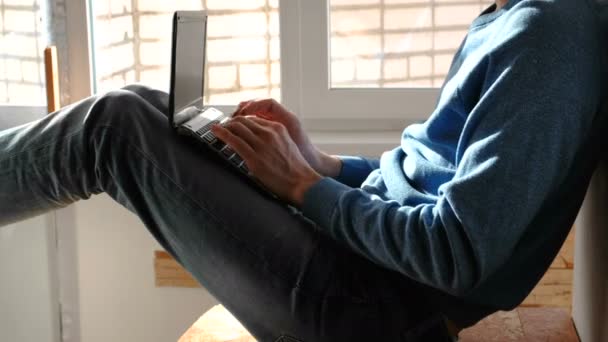 Εργάζονται για το laptop. Αγνώριστος νεαρός άνδρας που πληκτρολογείτε κάτι στο φορητό υπολογιστή του να κάθεται στο μπαλκόνι κοντά στο παράθυρο. — Αρχείο Βίντεο