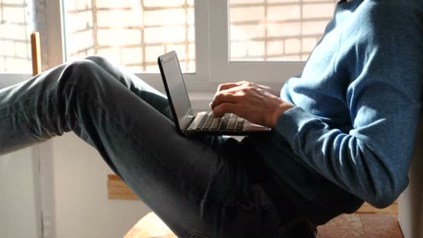 Arbeiten am Laptop. Unerkannter junger Mann tippt etwas in seinen Laptop, der auf dem Balkon am Fenster sitzt. — Stockvideo