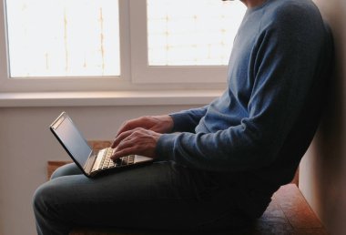 Dizüstü bilgisayar üzerinde çalışıyor. Diz üstü pencerenin yakınındaki balkonda oturan bir şey yazarak tanınmaz halde genç adam.