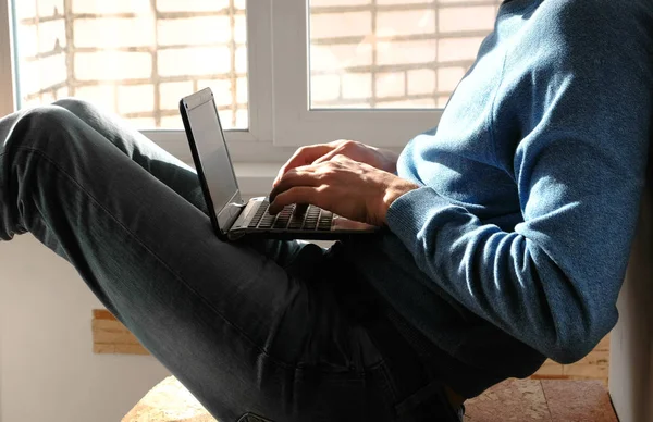 Dizüstü bilgisayar üzerinde çalışıyor. Diz üstü pencerenin yakınındaki balkonda oturan bir şey yazarak tanınmaz halde genç adam.