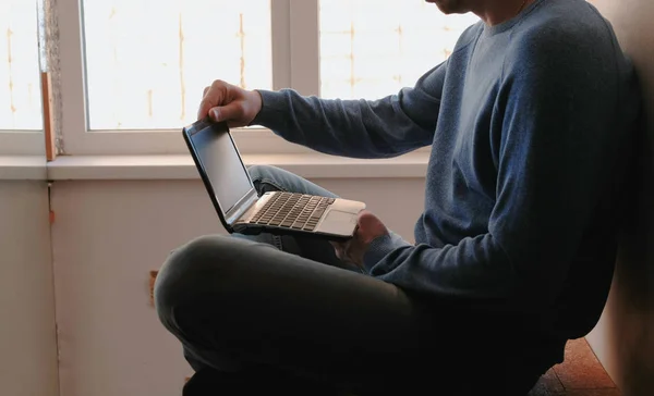 Работаю над ноутбуком. Неузнаваемый молодой человек открывает ноутбук и ждет, пока он загрузится, сидя на балконе возле окна . — стоковое фото