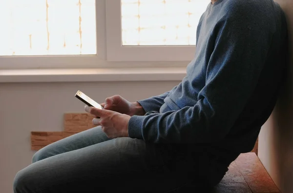 Человек с телефоном. Неузнаваемый молодой человек просматривает интернет-страницы в мобильном телефоне, сидя на балконе возле окна . — стоковое фото