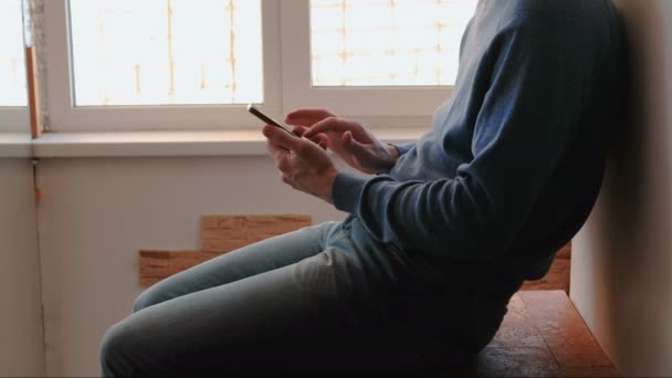 Man med telefonen. Oigenkännlig ung man chattar i mobiltelefon sitter på balkong nära fönstret — Stockvideo