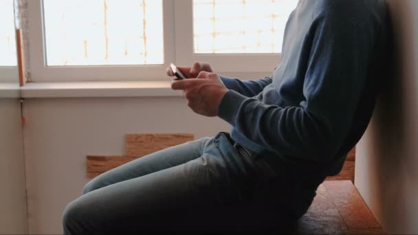 Un uomo con il telefono. Giovane irriconoscibile che gioca con il cellulare seduto sul balcone vicino alla finestra — Video Stock