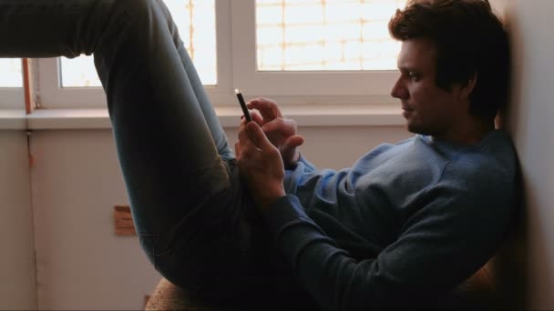 携帯電話を持つ男。若い男が窓の近くのバルコニーに座って携帯電話でチャット. — ストック動画