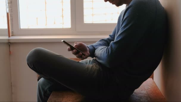 Man met telefoon. Onherkenbaar jongeman chatten in mobiele telefoon zit op balkon dichtbij het venster. — Stockvideo