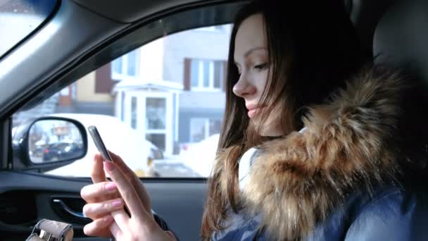 Frau im Auto mit Handy. junge schöne brünette Frau schaut etwas in das Handy und wendet sich der Kamera zu. — Stockvideo