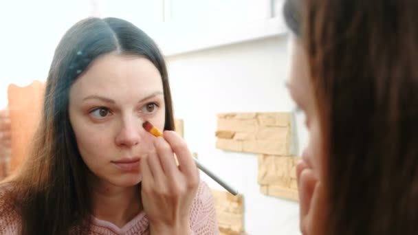 Mujer pone una crema tonal Fundación en la cara usando un cepillo delante del espejo . — Vídeos de Stock