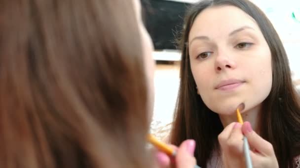 Mulher coloca um creme tonal Foundation no rosto usando um pincel na frente do espelho. Fechar as mulheres vista frontal . — Vídeo de Stock